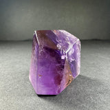 Polished ametrine crystal standing on a gray surface, displaying a gradient of purple and golden-yellow hues with natural internal fractures and angular facets.