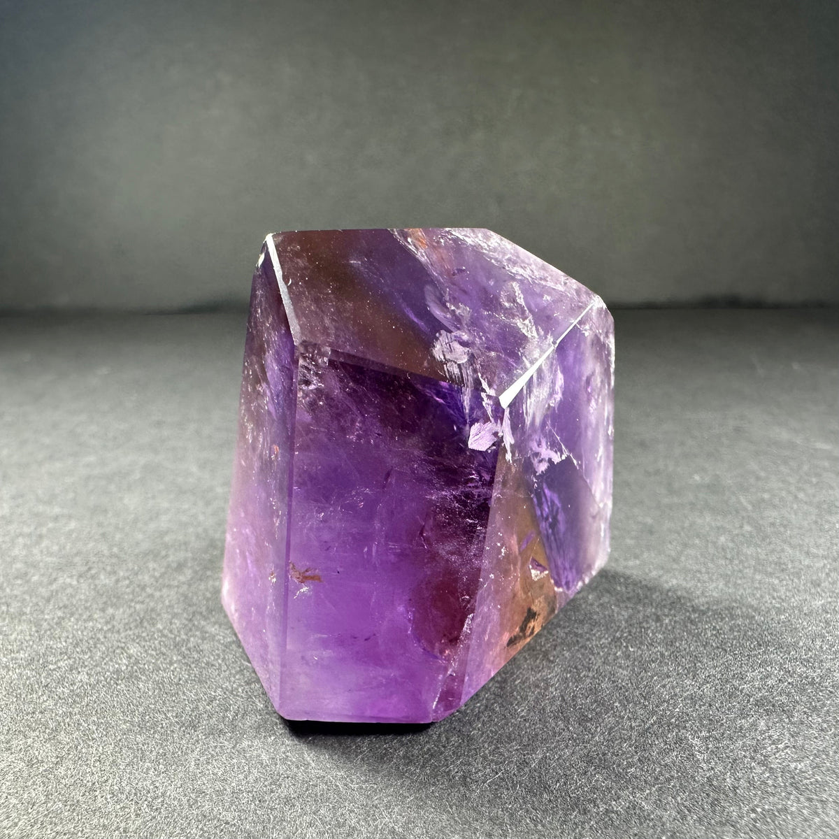 Polished ametrine crystal standing on a gray surface, displaying a gradient of purple and golden-yellow hues with natural internal fractures and angular facets.