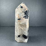 Tapered tower-shaped peach moonstone crystal with translucent white base and scattered black inclusions, standing on a gray surface against a gray background.