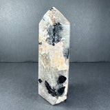Peach moonstone tower from Brazil with a pointed tip, showing a pale peach and white base with scattered black mineral patches on a dark gray background.