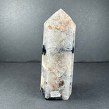 Tapered translucent peach moonstone tower with black and white inclusions standing upright on a gray surface against a gray background.