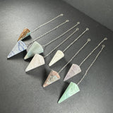 Seven faceted stone pendulums in various colors including blue, green, pink, and white, each attached to a silver chain laid out diagonally on a dark surface.