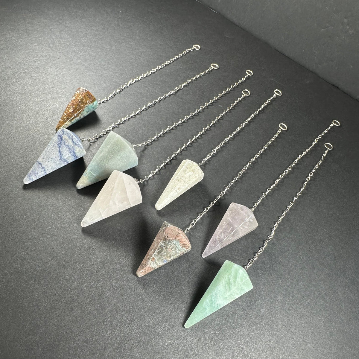 Seven faceted stone pendulums in various colors including blue, green, pink, and white, each attached to a silver chain laid out diagonally on a dark surface.