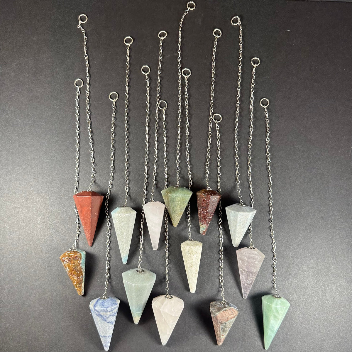 Thirteen long-chain pendulums with faceted stone weights in various earth tones and pastel colors, arranged vertically on a dark background.