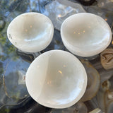 Three translucent white selenite bowls with smooth rounded shapes, displayed from above on a glossy agate slab with swirls of grey and beige.