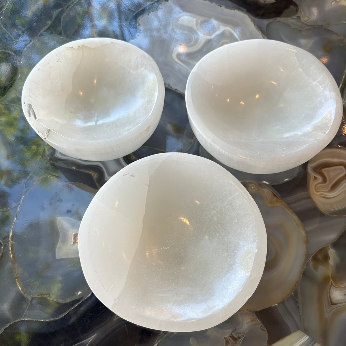 Three translucent white selenite bowls with smooth rounded shapes, displayed from above on a glossy agate slab with swirls of grey and beige.