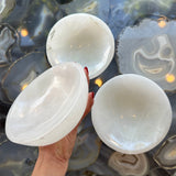 Hand holding one translucent white selenite bowl with smooth, rounded edges above two similar bowls on a polished agate slab surface with circular, layered patterns in blue and gray tones.