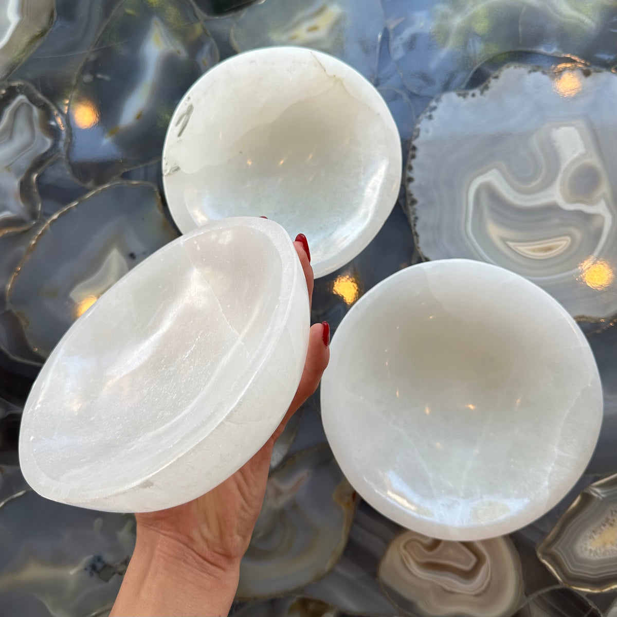Hand holding one translucent white selenite bowl with smooth, rounded edges above two similar bowls on a polished agate slab surface with circular, layered patterns in blue and gray tones.