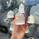 Hand holding a translucent white selenite obelisk with a pointed top, surrounded by similar obelisks on a polished gray and black agate surface.