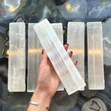 Five long, translucent white selenite bars with fibrous texture, one held by a hand with red nails above a dark polished stone background with swirling patterns.
