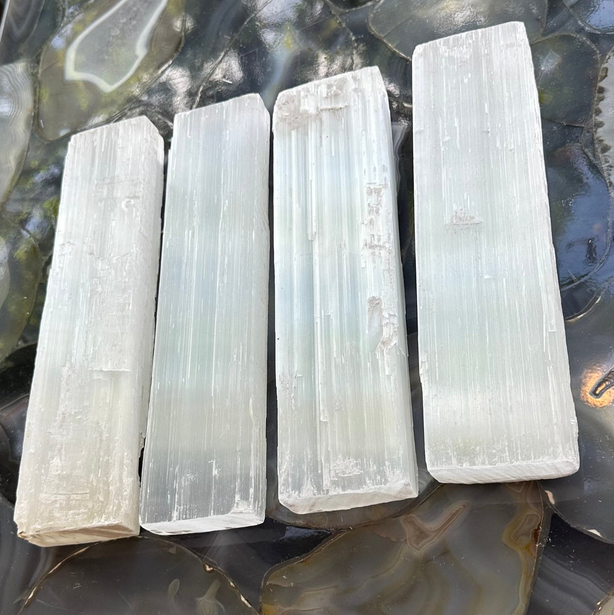 Four elongated rough selenite bars with vertical striations and a translucent white-gray sheen, arranged side by side on a glossy, multicolored agate surface.