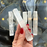 Hand holding a translucent white selenite stick with a fibrous texture above five similar sticks on a glossy black and gray agate stone surface with yellow reflections.