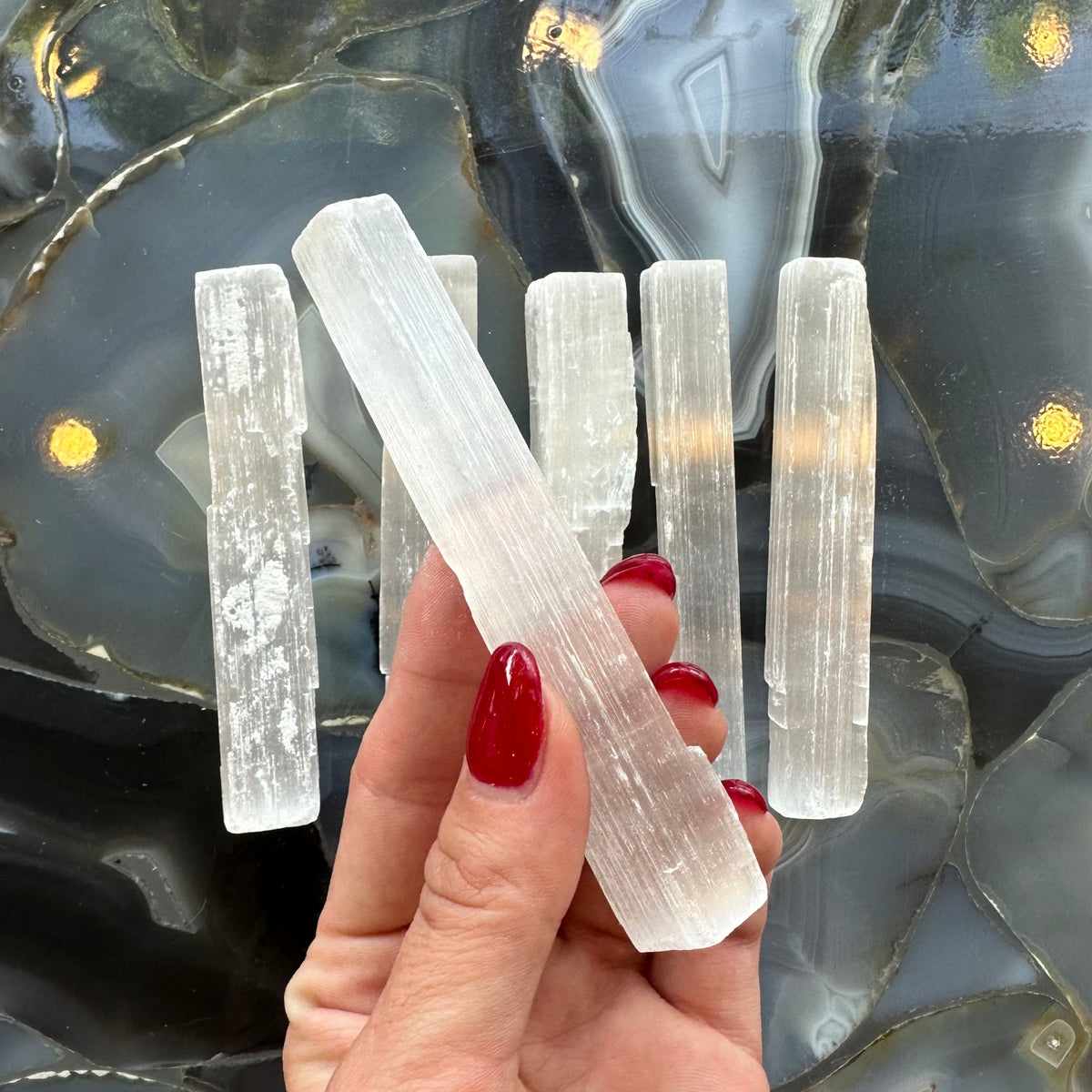Hand holding a translucent white selenite stick with a fibrous texture above five similar sticks on a glossy black and gray agate stone surface with yellow reflections.