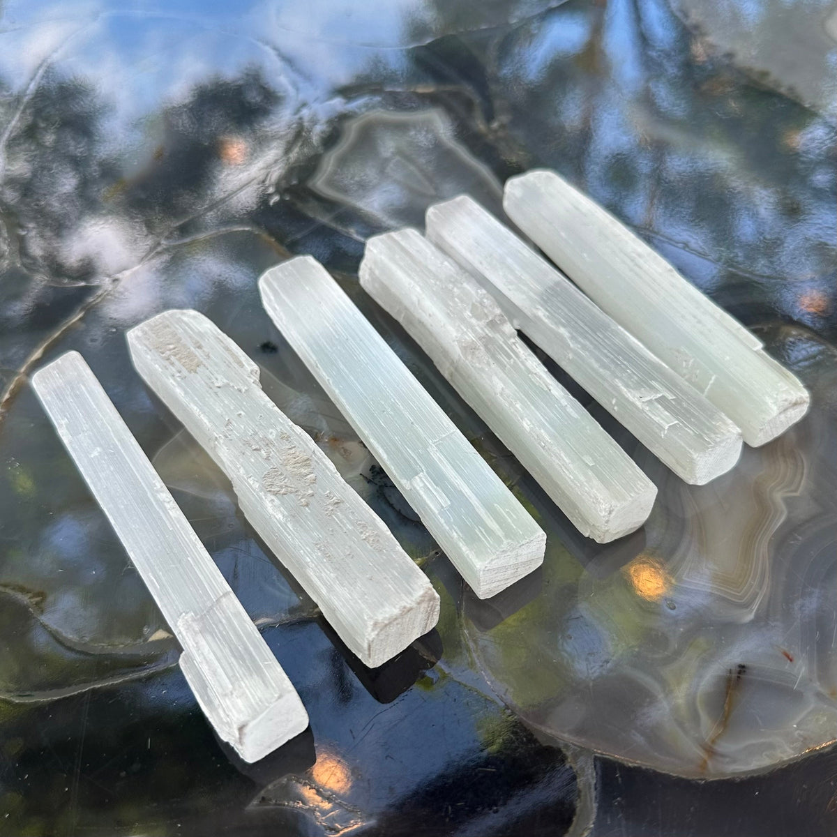 Six translucent, elongated white selenite sticks, each about 15cm long, displayed on a polished agate slice with blue and gray tones and natural banding patterns.