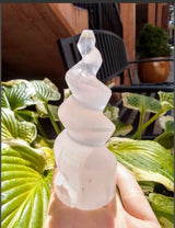 Hand holding a translucent white selenite spiral tower with a twisting, conical shape, set against green leaves and outdoor patio furniture in the background.