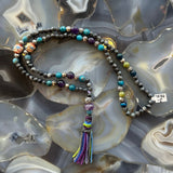 Beaded mala necklace with a mix of larvikite and colorful stone beads, featuring a multicolored thread tassel, displayed on a polished agate surface with swirling gray and cream patterns.