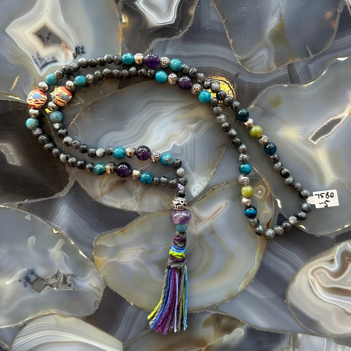 Beaded mala necklace with a mix of larvikite and colorful stone beads, featuring a multicolored thread tassel, displayed on a polished agate surface with swirling gray and cream patterns.