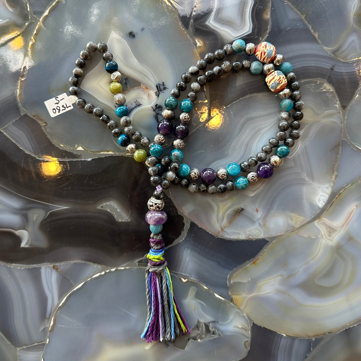 Mala necklace with mixed stone beads including larvikite, turquoise, purple, and earth tones, finished with a multicolored tassel, displayed on a polished agate surface.