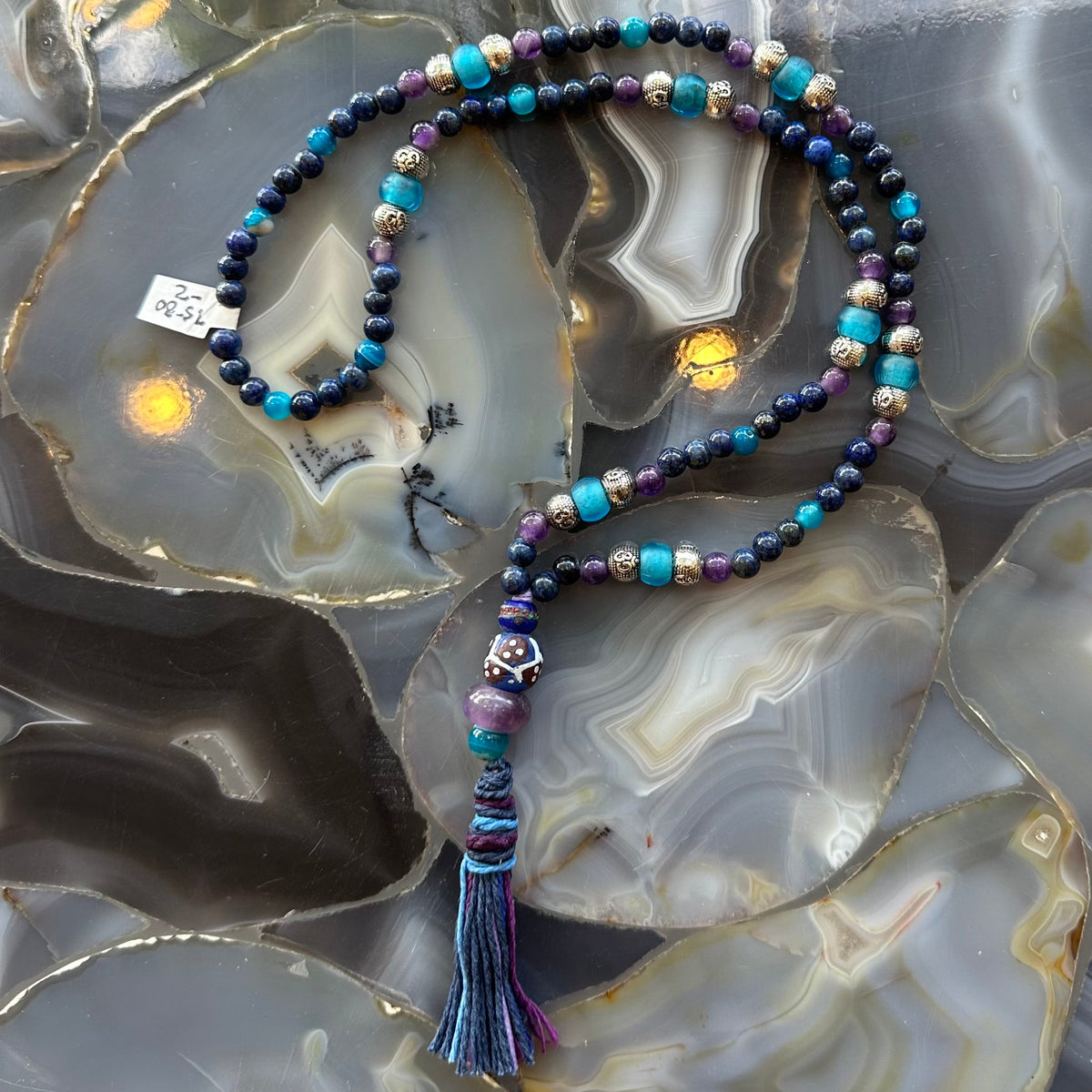 Lapis lazuli mala with deep blue, turquoise, purple beads, silver spacers, and a multicolored thread tassel laid on a polished agate slab with layered gray and amber patterns.