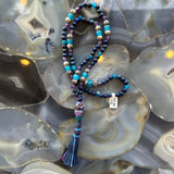 Lapis lazuli mala necklace with mixed blue, purple, turquoise, and white beads, featuring a purple pendant and blue tassel, displayed on a gray and beige agate surface.