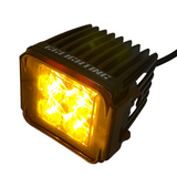 Compact black finned LED pod light with glowing amber lens cover and white logo on top, angled view showing cable extending from back.