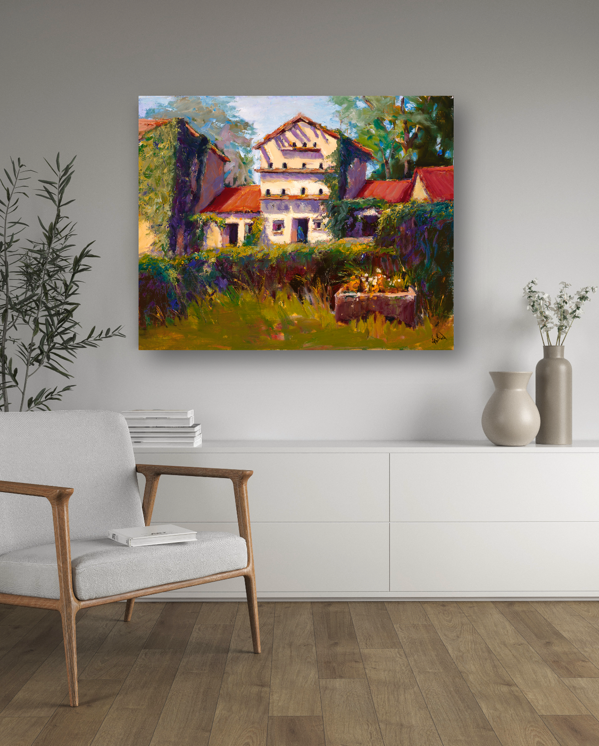 Impressionist painting of a white house with red roofs surrounded by greenery and a small stone structure in the foreground, displayed on a white wall above a modern sideboard.