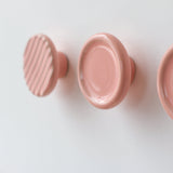 Three round pink resin coat hooks mounted on a white wall, with the leftmost hook featuring diagonal ridges and the middle hook a smooth concave design.