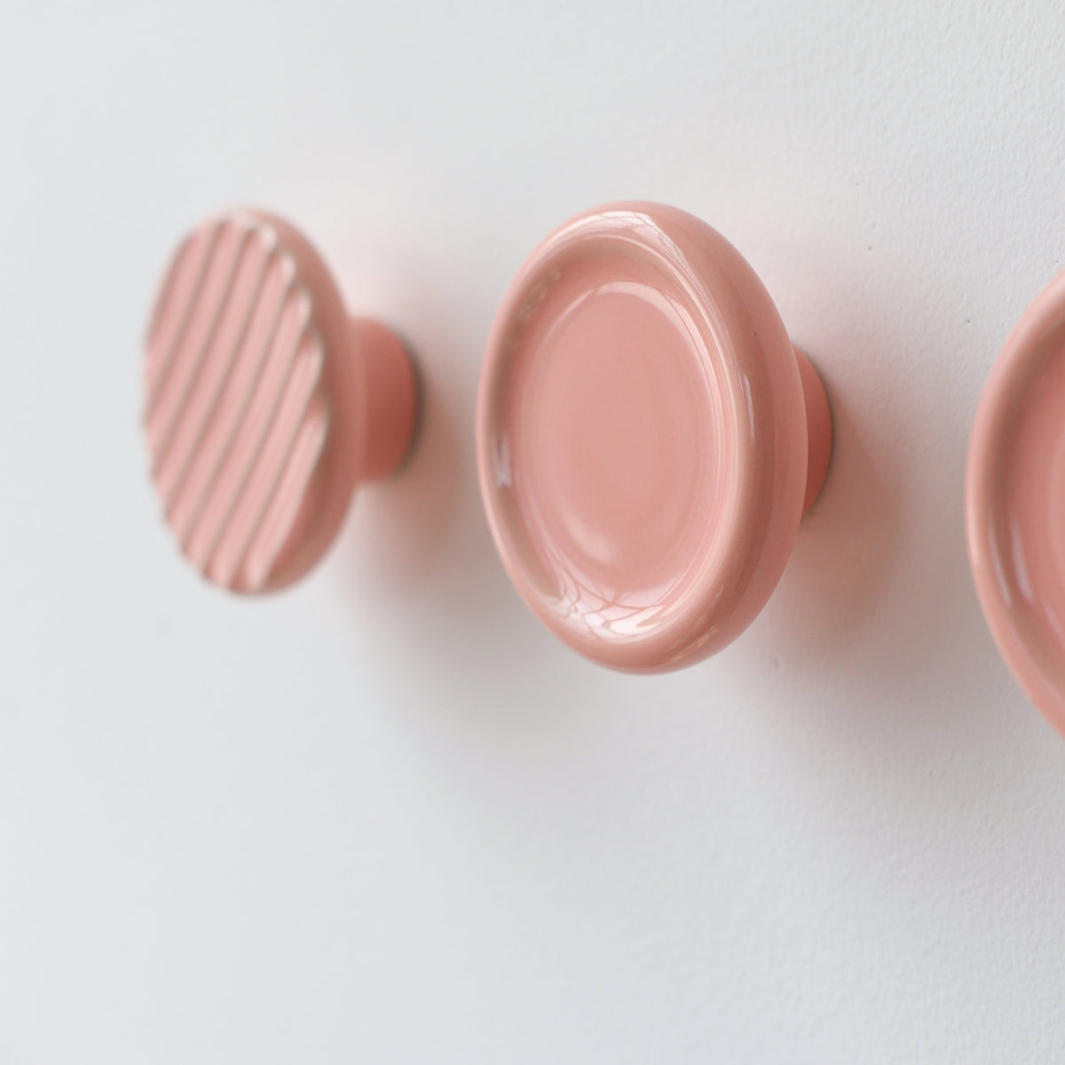 Three round pink resin coat hooks mounted on a white wall, with the leftmost hook featuring diagonal ridges and the middle hook a smooth concave design.