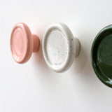Three round, glossy coat hooks mounted on a white wall, colored pink, white with multicolored specks, and dark green, shown from a side angled view.