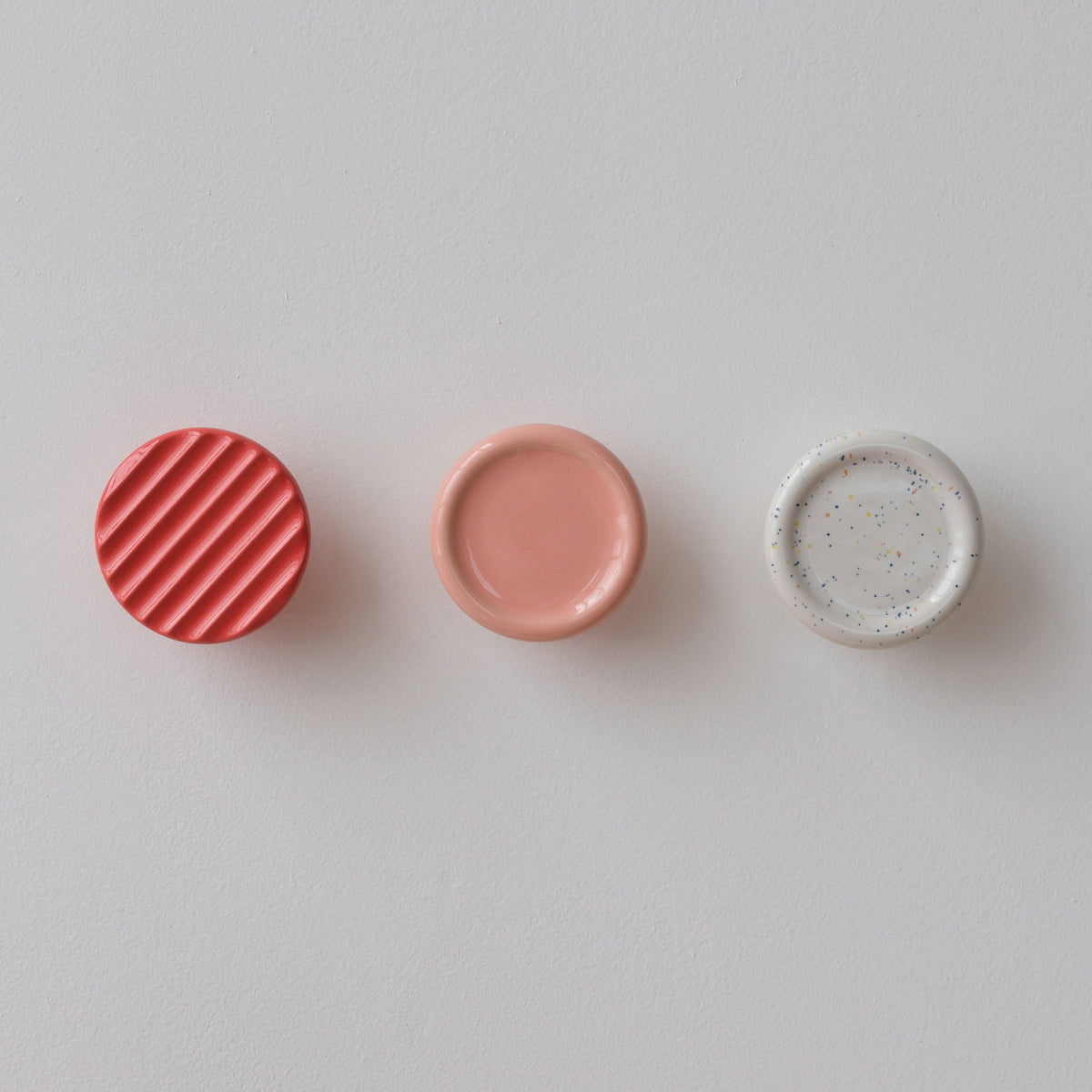 Three round coat hooks on a white wall, from left: red with diagonal ridges, smooth muted pink, and white speckled with small colorful dots.