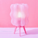 Pink wavy lamp with a translucent ribbed shade and three slender legs, standing against a matching pink backdrop on a pale pink surface.
