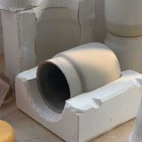 Gray ceramic coffee tumbler mug in a white plaster mold with visible edges and casting details on a wooden surface under soft light.