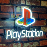 Illuminated neon sign of the PlayStation logo with red, blue, yellow, and green colors above glowing white 'PlayStation' text on a brick wall background.