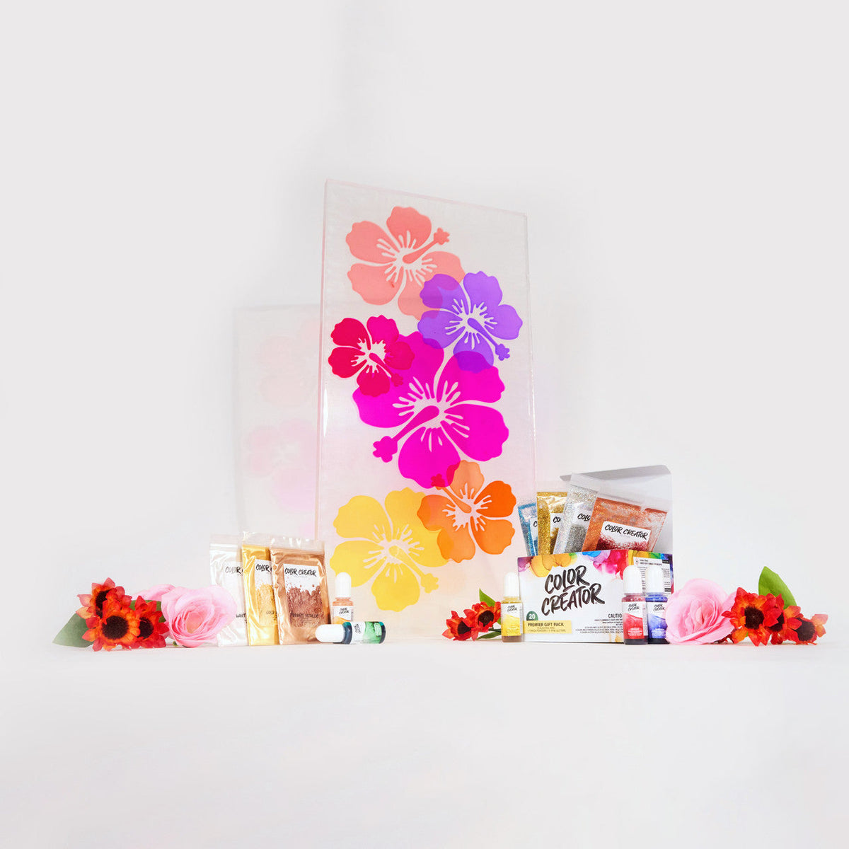 A platter decorated with vibrant pink, purple, orange, and yellow hibiscus flowers flanked by resin pigment powders, color and mica powders, and artificial red and pink flowers on a white background.