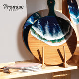 Round wooden paddle board with blue, green, and white ocean wave resin art on one side, displayed on a wooden rack with sunlight casting shadows.