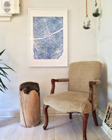Beige upholstered wooden armchair with curved legs beside a natural wood stump stool, under a framed blue tree ring print on a white wall with hanging planters.