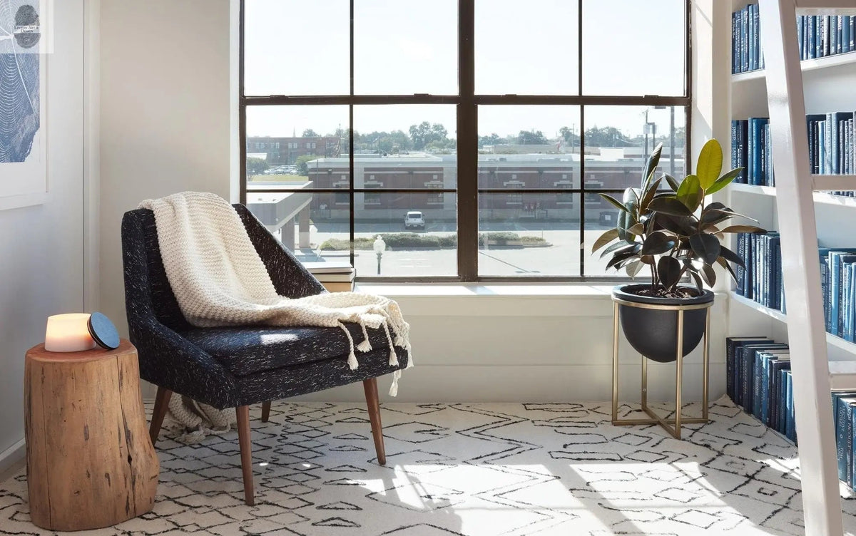 Cozy corner with a dark cushioned chair draped in a white knit blanket, natural wood stump side table, and a large window overlooking an urban landscape.