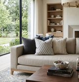Beige sofa with five decorative pillows in ash grey, patterned grey, blue with white floral, tan textured, and beige, in a living room with wooden shelves and large window.