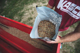 Person holding an open bag filled with mixed seeds or pellets over a red feeder partially filled with the same mix, outdoors on grassy ground.