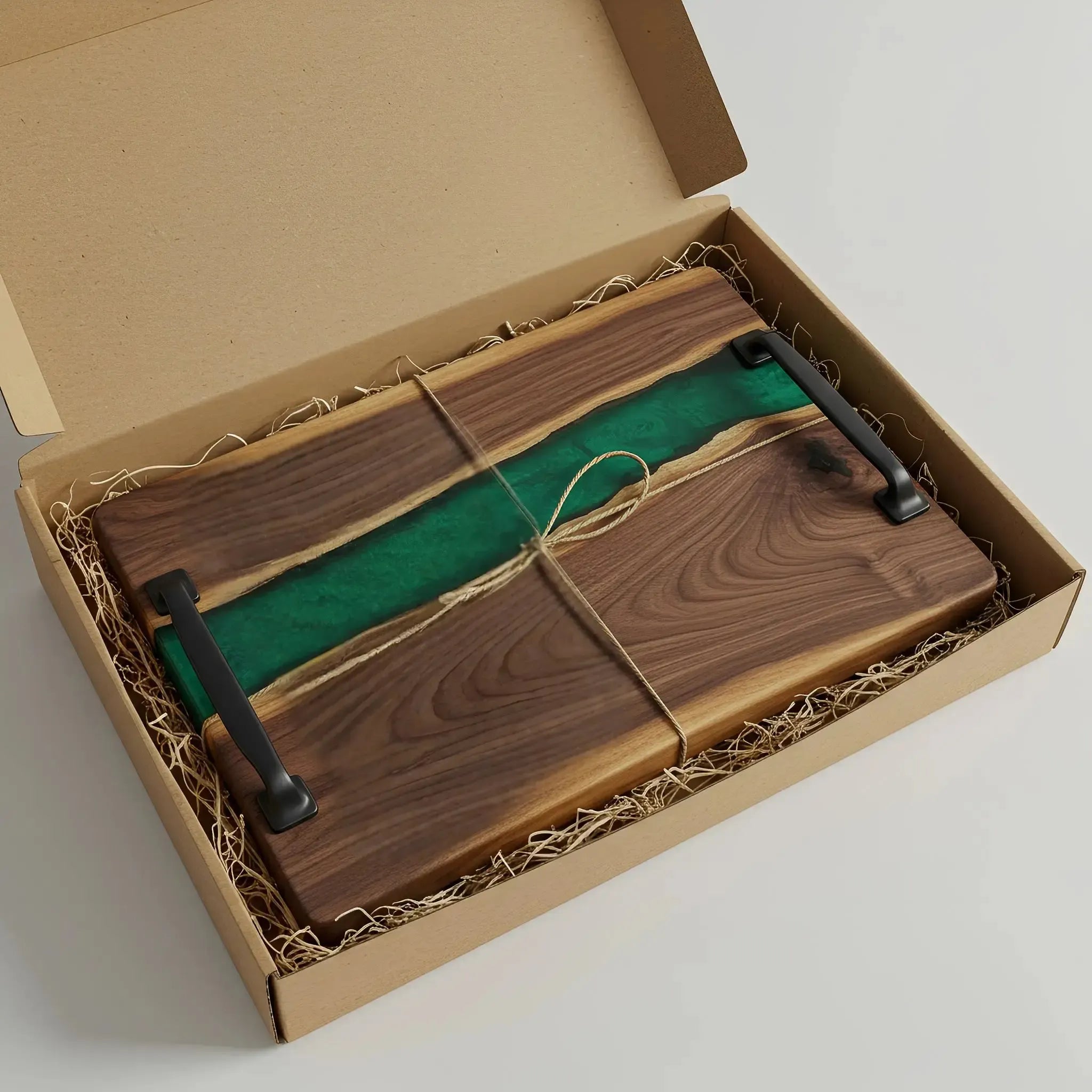 a charcuterie board carefully packaged for shipping. The board is made from live edge black walnut with a smooth, polished finish and a emerald green epoxy river running through the center. It’s wrapped in kraft paper and tied neatly with natural twine for a handmade, artisan feel. Matte black metal handles are attached at both ends