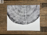 Black ink print of pine tree rings on white paper, centered and cropped horizontally on rustic wooden surface with varied wood grain and color.