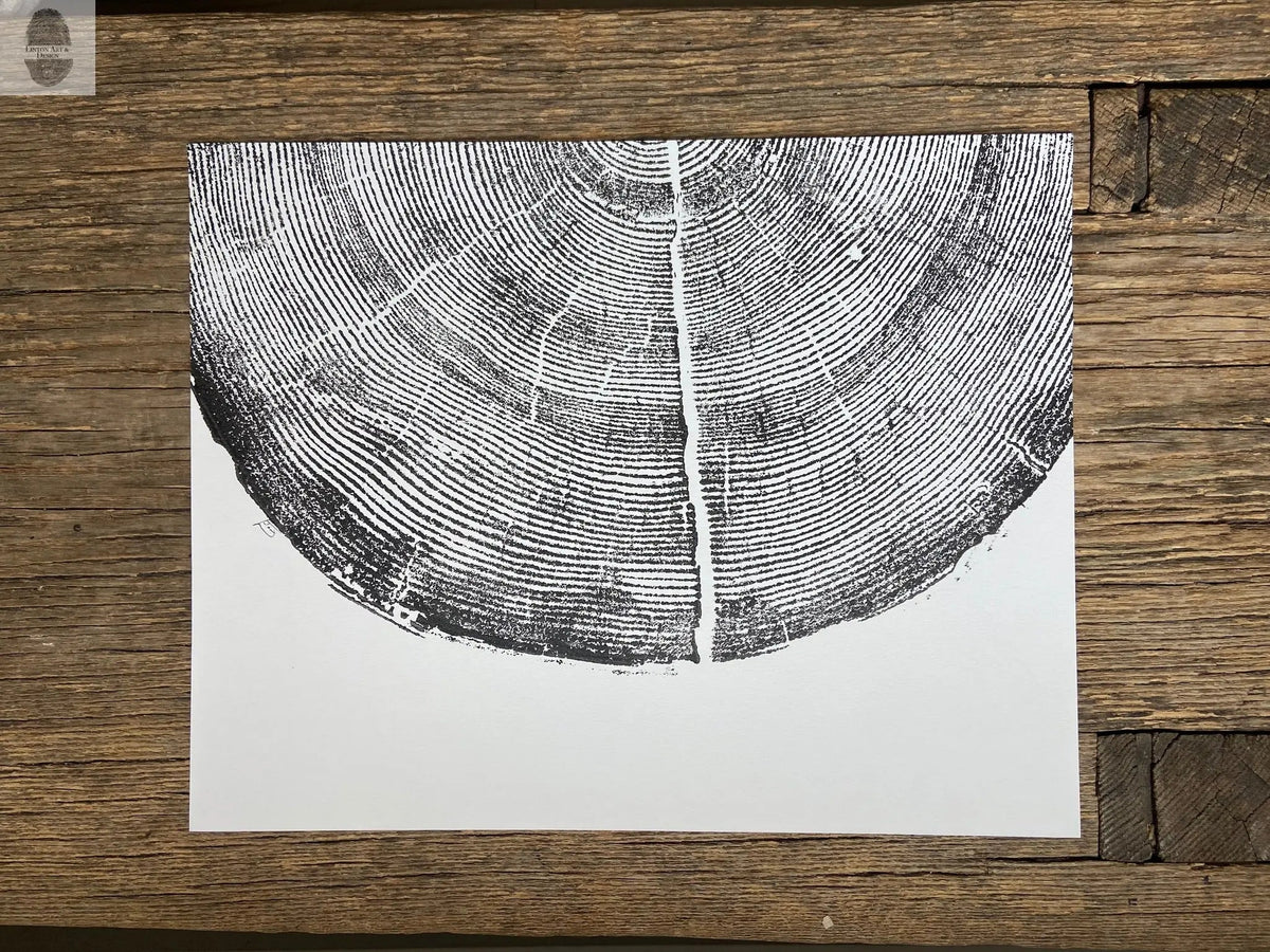 Black ink print of pine tree rings on white paper, centered and cropped horizontally on rustic wooden surface with varied wood grain and color.