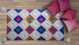 Rectangular woven rug with colorful diamond pattern in vibrant pink, blue, orange, and white fluffy textured squares, bordered by tassels, with three pink pillows on light wood floor