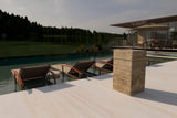 Square travertine marble side table with LED illumination on a light stone patio beside an infinity pool overlooking a wooded lake at dusk.