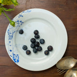 White ceramic plate with blue hand-drawn floral and geometric accents, holding scattered blueberries, on a dark wooden surface next to vintage silver spoons.