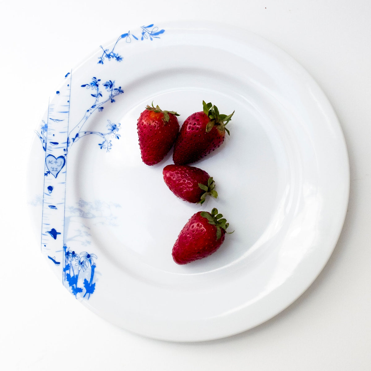 White round plate with blue hand-drawn birch tree and foliage design on the rim, holding four red strawberries with green leaves at center.