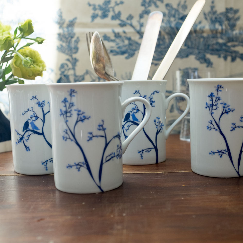 Set of white mugs with blue illustrations of birds perched on delicate branches, arranged on a wooden table with cutlery inside one mug and a green flower nearby.