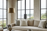 Neutral-toned living room with natural light, featuring a beige sofa adorned with scalloped textured woven pillows, tall brass floor lamp, and large window overlooking greenery.
