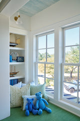 Cozy window nook with green carpet, featuring two blue teddy bears and patterned green pillows beside white-framed double windows overlooking trees and a parked car.
