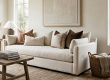 Cream-colored sofa with brown and beige pillows, including a Silt Brown District Pillow Cover, positioned next to a woven basket and wooden coffee table with stacked books.